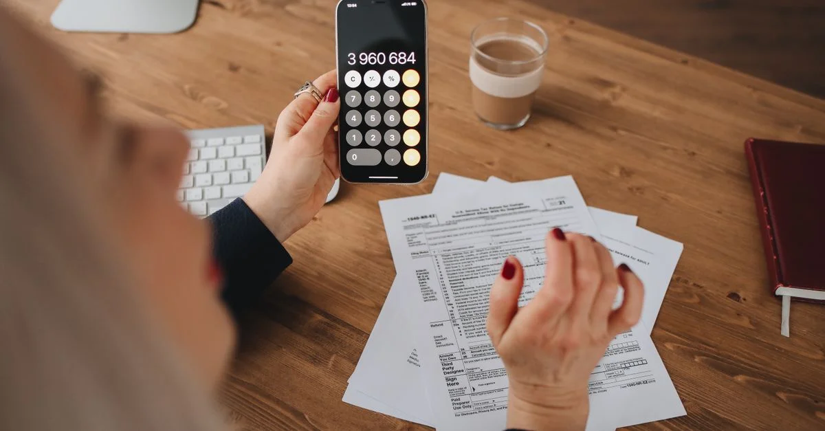 Une femme calcule sur son smartphone tout en travaillant avec des documents à un bureau.
