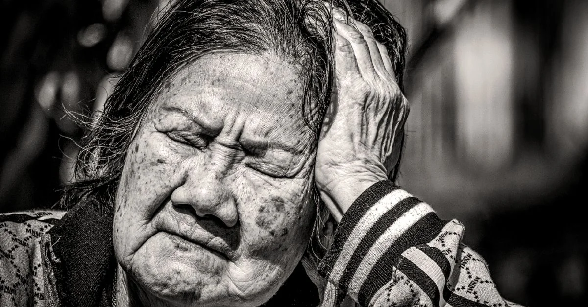 Gros plan sur une femme âgée les yeux fermés, assise à l'extérieur en noir et blanc.