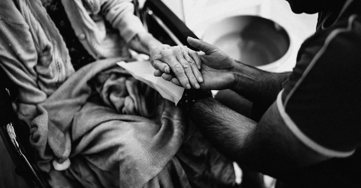 Un aidant tenant les mains d'une femme âgée dans un fauteuil roulant, soulignant l'empathie et le soutien.