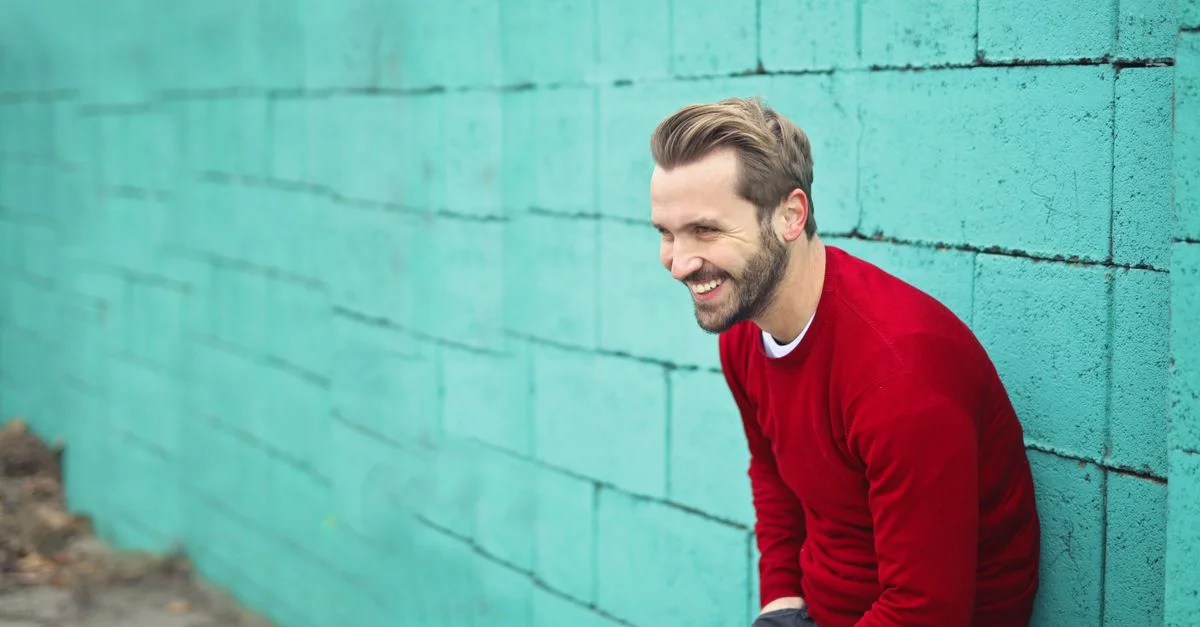 Homme souriant à Budapest portant un pull rouge contre un mur en briques teal, dégageant du bonheur.