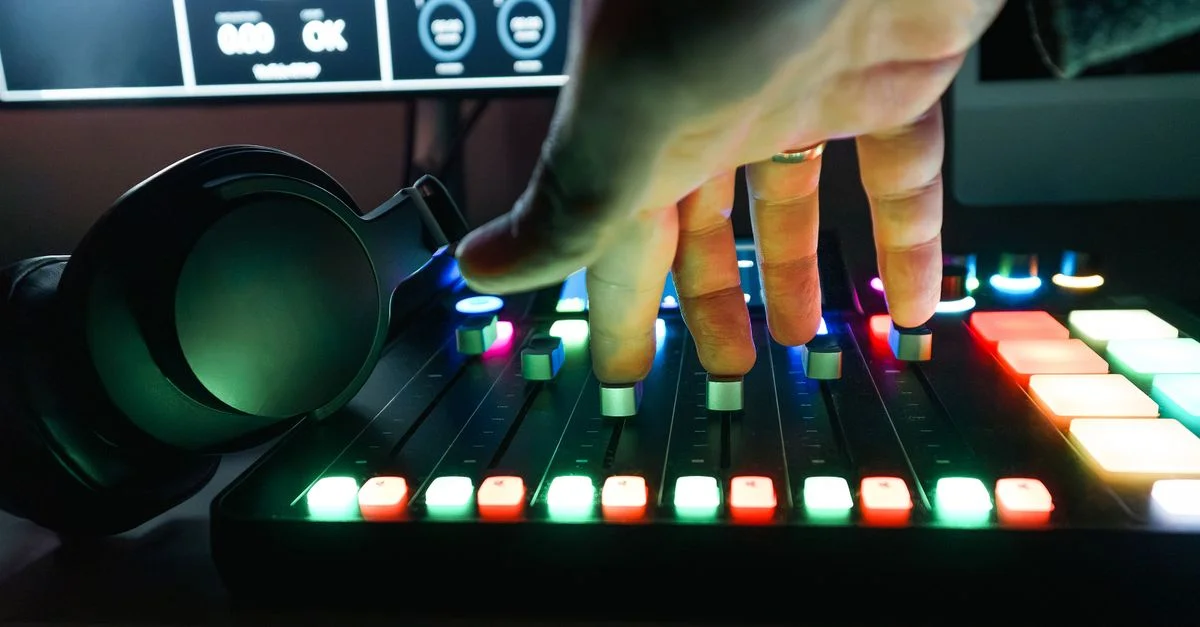 Gros plan sur une main ajustant les curseurs d'une console de mixage audio colorée, avec un casque audio à proximité dans un studio d'enregistrement.