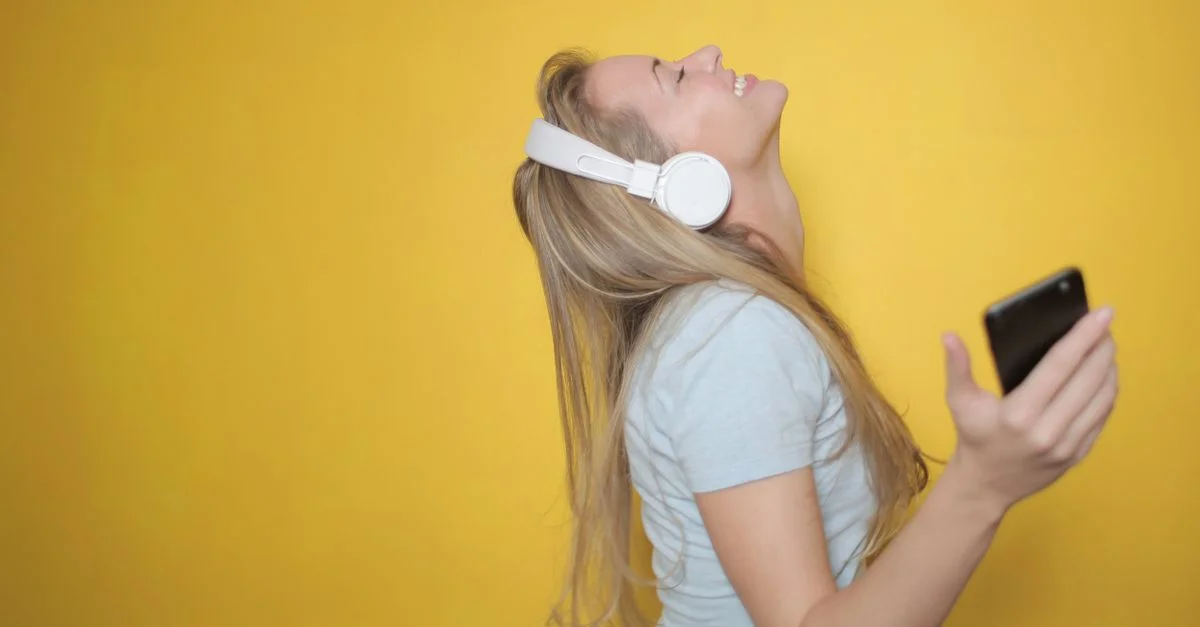 Femme blonde profitant de la musique avec des écouteurs et un smartphone sur un fond jaune.