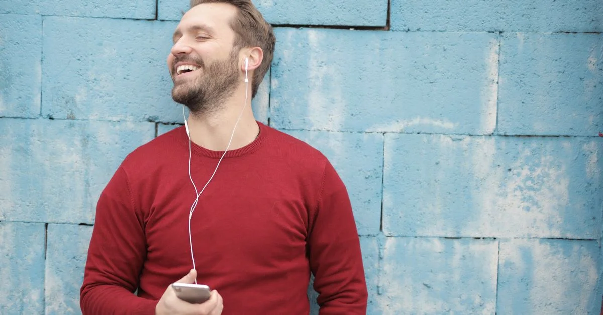 Homme heureux portant des écouteurs, souriant devant un mur bleu tout en écoutant de la musique en extérieur.